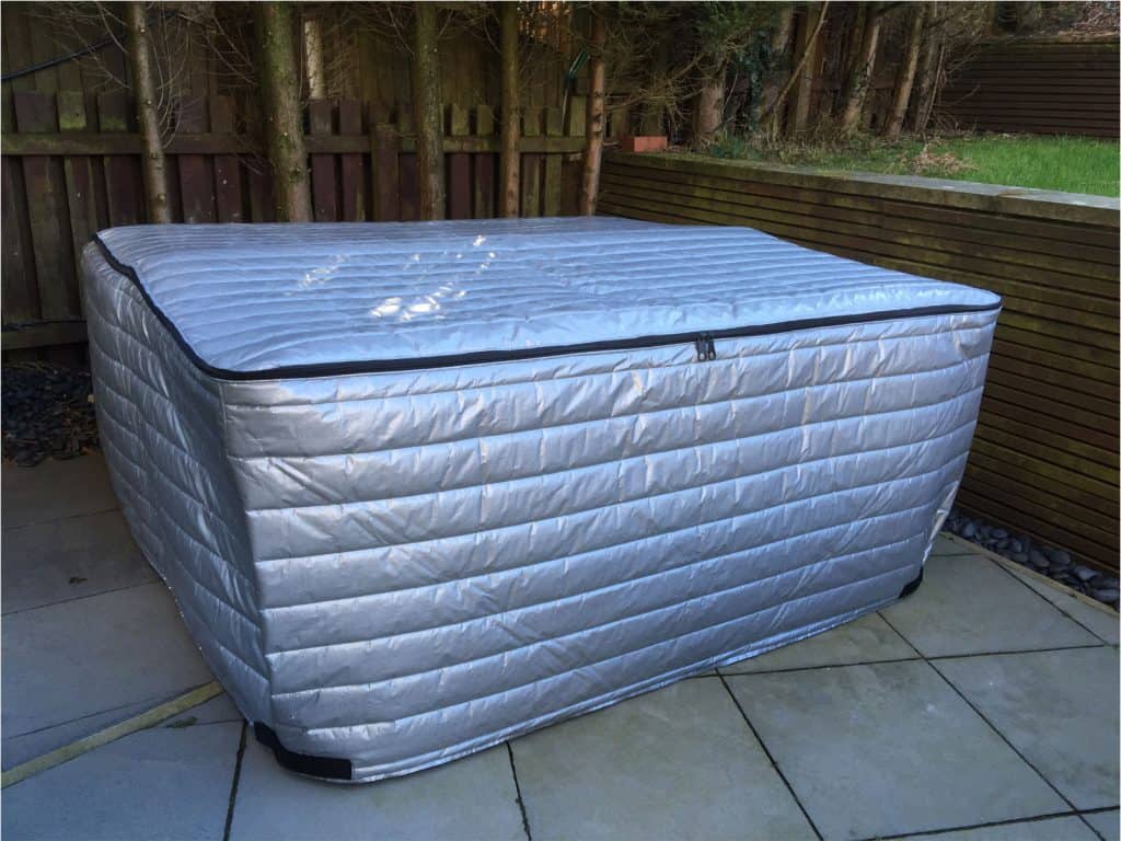 A hot tub fitted with the Deluxe Insulated hot tub cover is situated on a tiled patio adjacent to a wooden fence and grassy area.